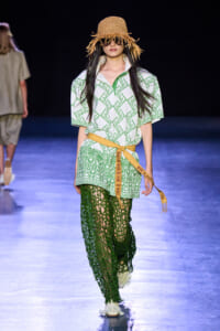 Model walks the runway in a white shirt with a green geometric print, a tan belt, and emerald crochet pants, wearing a straw hat and sunglasses.