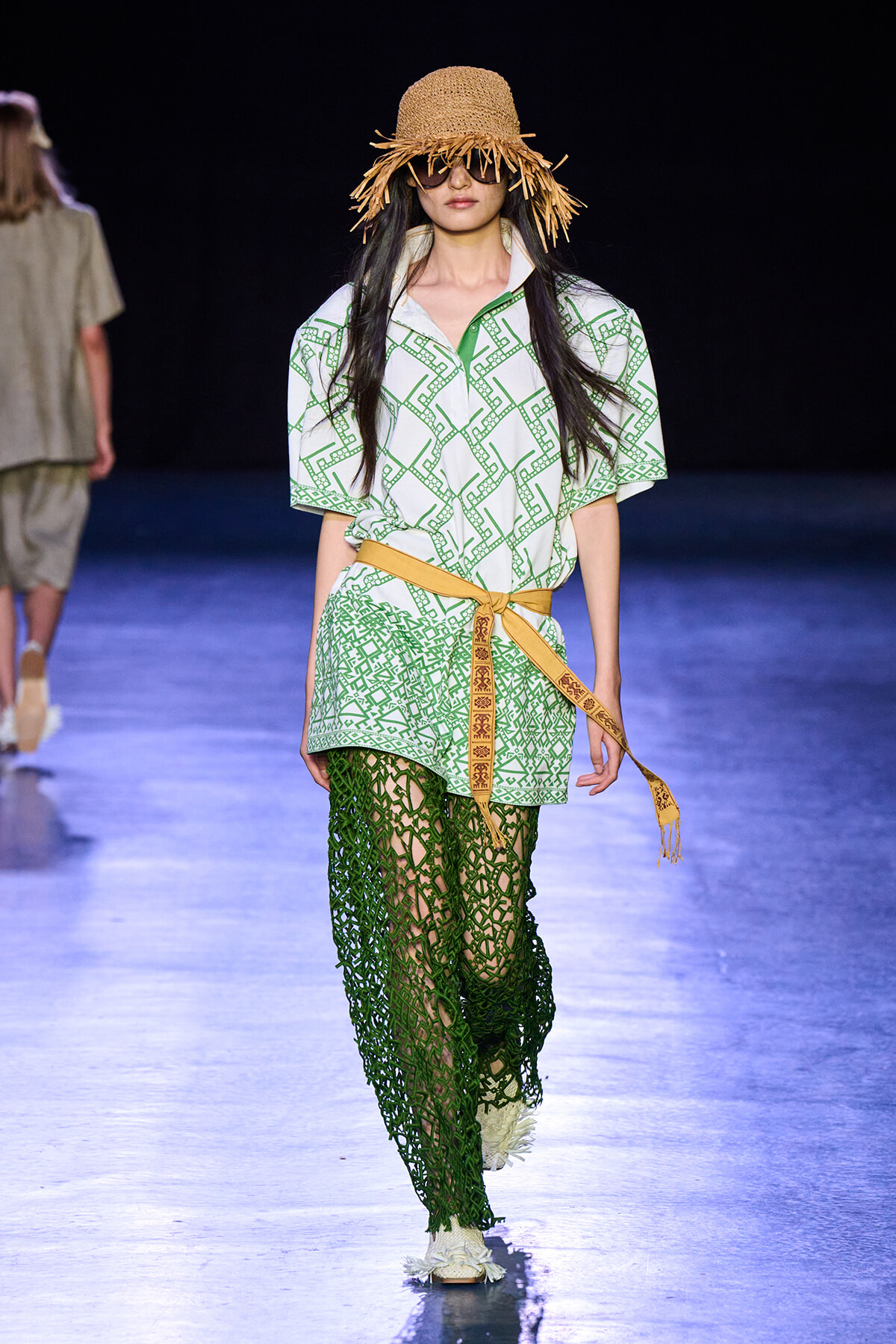 Model walks the runway in a white shirt with a green geometric print, a tan belt, and emerald crochet pants, wearing a straw hat and sunglasses.