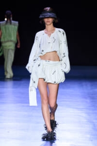 Model walks the runway in a white cropped blouse and matching high-waisted shorts/skirt with black abstract marks; oversized black visor hat and feathered footwear.