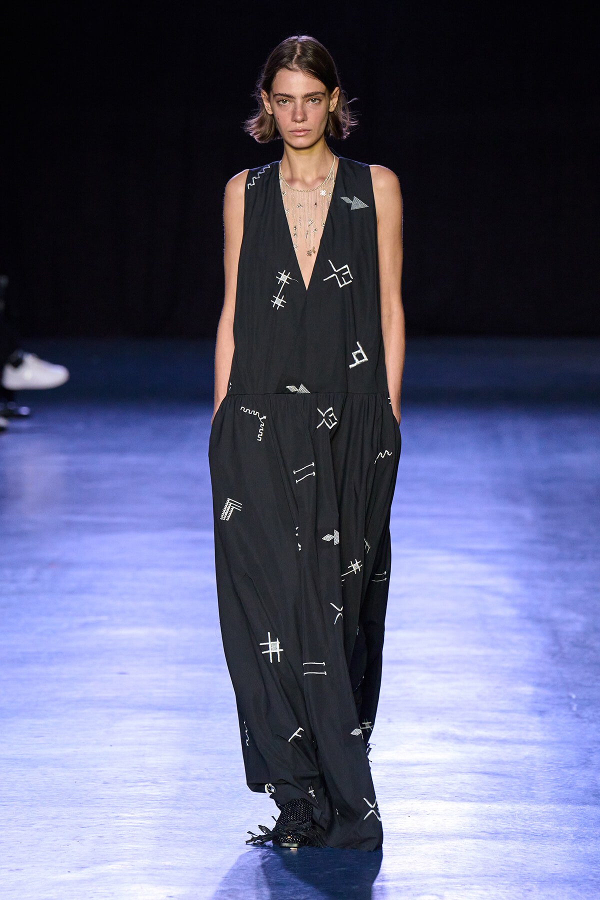 Model walking a runway in a loose black jumpsuit with white stitched symbols against a dark backdrop.