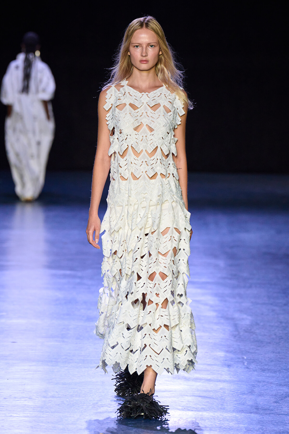 Model walking a runway in a white lattice dress with geometric cutouts and black feathered shoes on a dark backdrop.