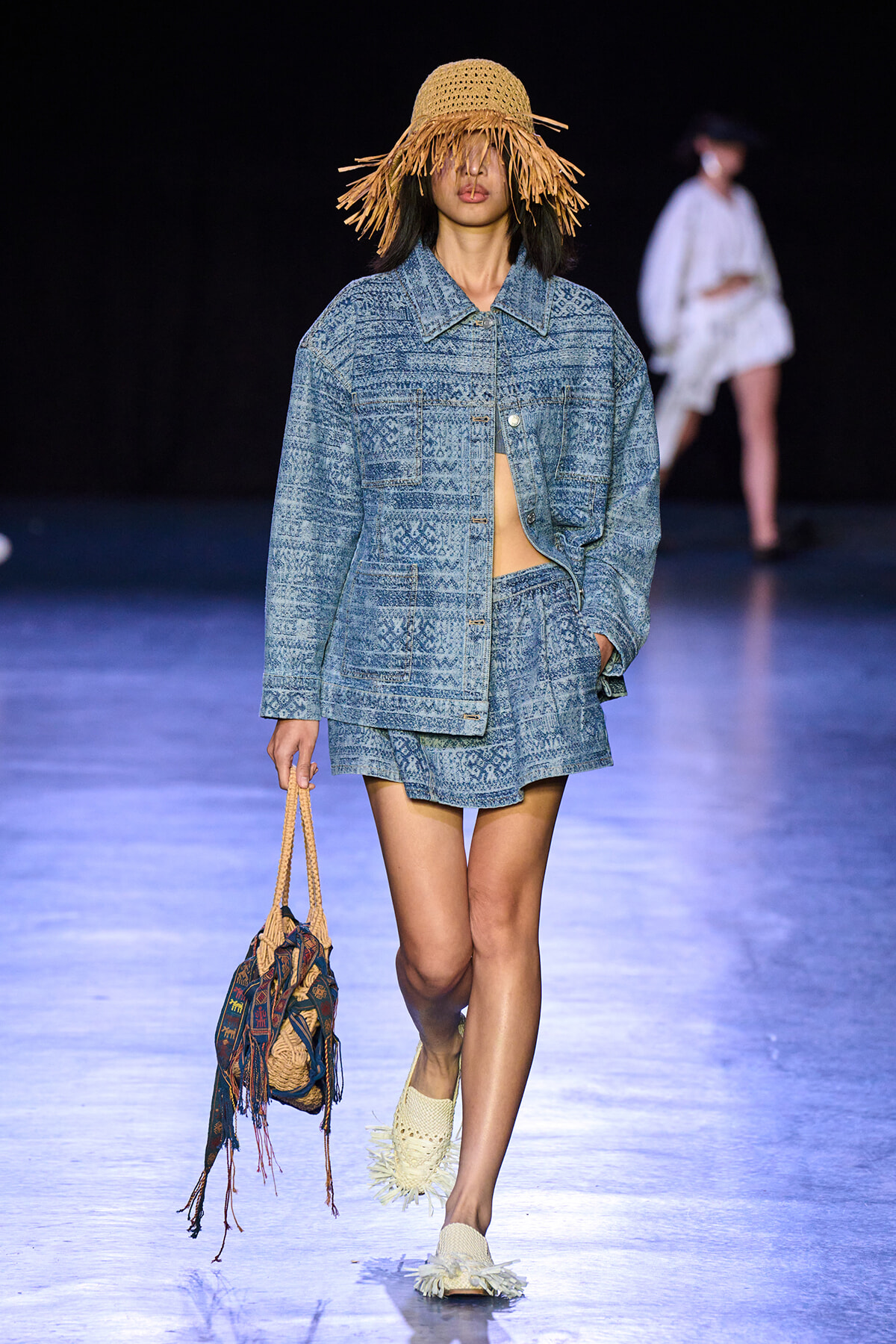 Model walking a fashion runway in a blue patterned denim suit with a matching jacket and shorts, wearing a wide woven straw hat and carrying a fringed patterned bag.