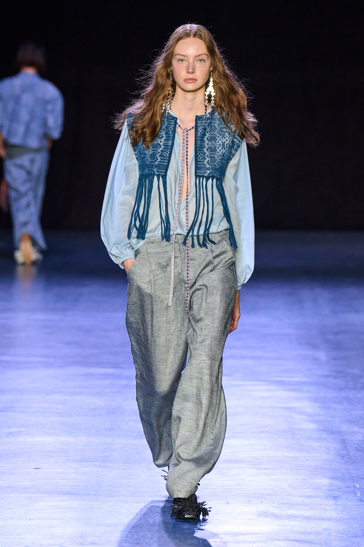Model walks the runway in a blue embroidered fringe vest over a pale blue blouse and gray loose trousers, with long geometric earrings catching the lights.