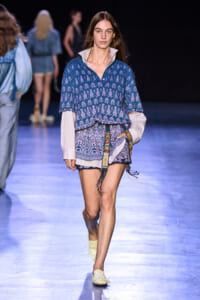 Model walks the runway wearing a blue patterned blouse layered over a pale striped shirt and matching shorts, with a decorative belt and cream woven shoes.