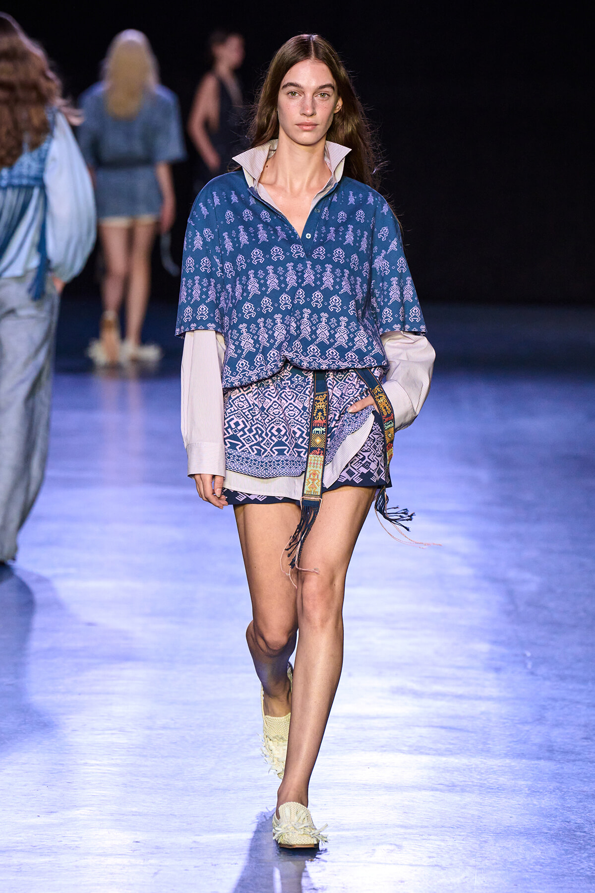 Model walks the runway wearing a blue patterned blouse layered over a pale striped shirt and matching shorts, with a decorative belt and cream woven shoes.