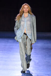 Female model walks down a fashion runway in a loose gray suit with a patterned sheer shirt and a decorative belt, against a dark backdrop.