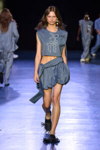 Model walks the runway in a blue denim two-piece: cropped sleeveless top with white geometric embroidery and a voluminous bubble skirt, with statement earrings.