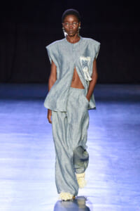 Full-body shot of a dark-skinned model walking a runway in a light blue, loose two-piece linen outfit with a fringed pocket patch and tassel accents at the hems.