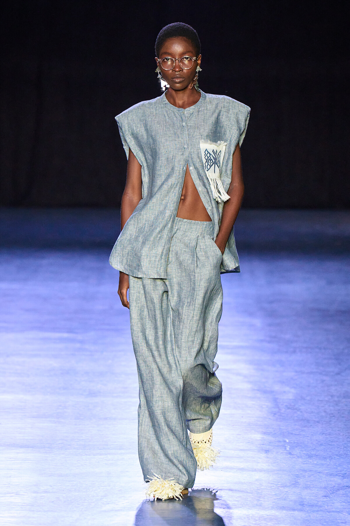 Full-body shot of a dark-skinned model walking a runway in a light blue, loose two-piece linen outfit with a fringed pocket patch and tassel accents at the hems.