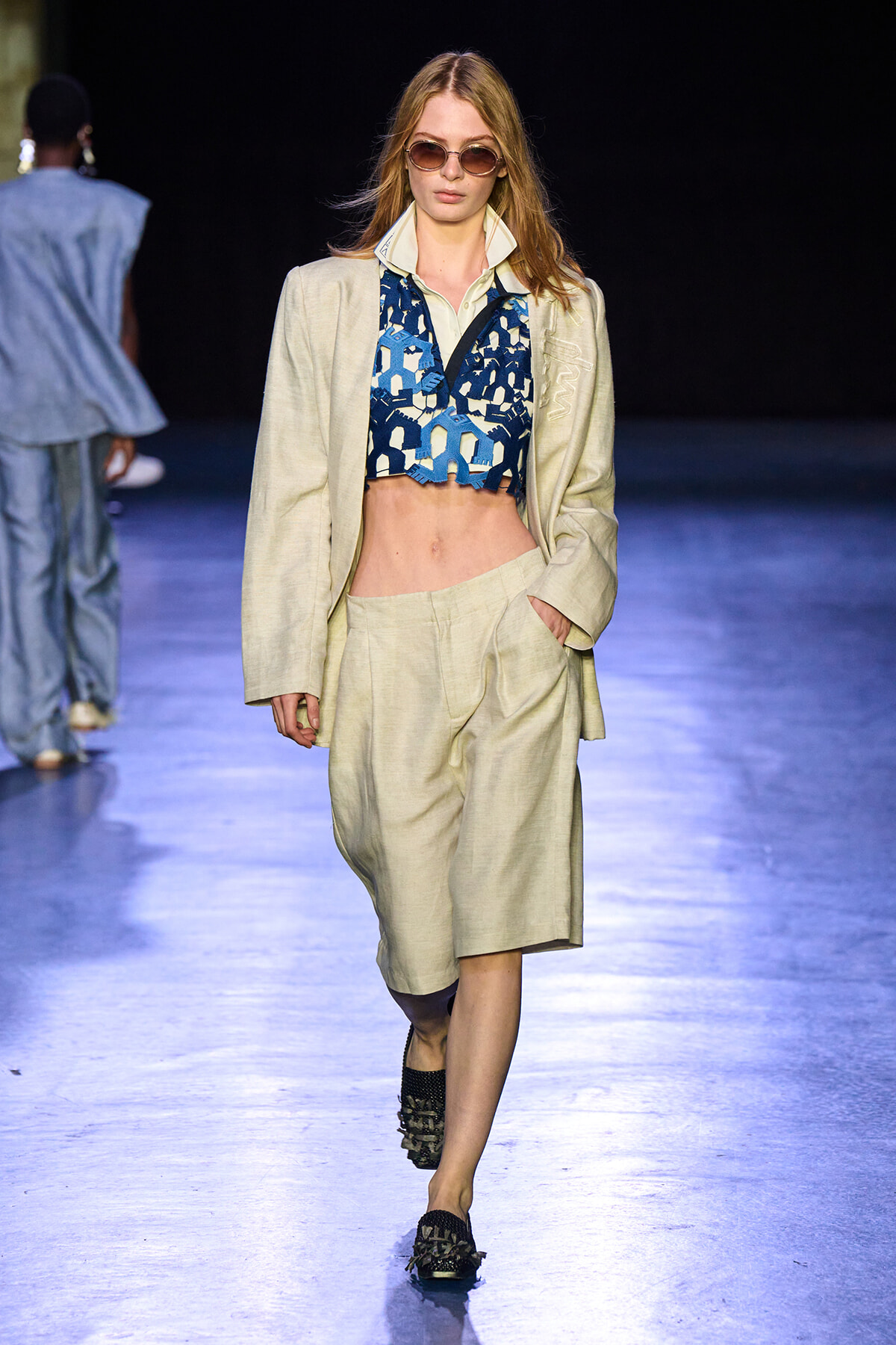 Model walks the runway in a beige suit with a blue geometric crop top and round sunglasses under bright lights.