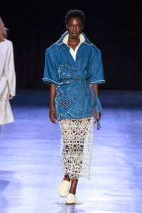 Runway model wears a blue denim tunic with geometric embroidery, paired with a white crochet skirt and light woven shoes.