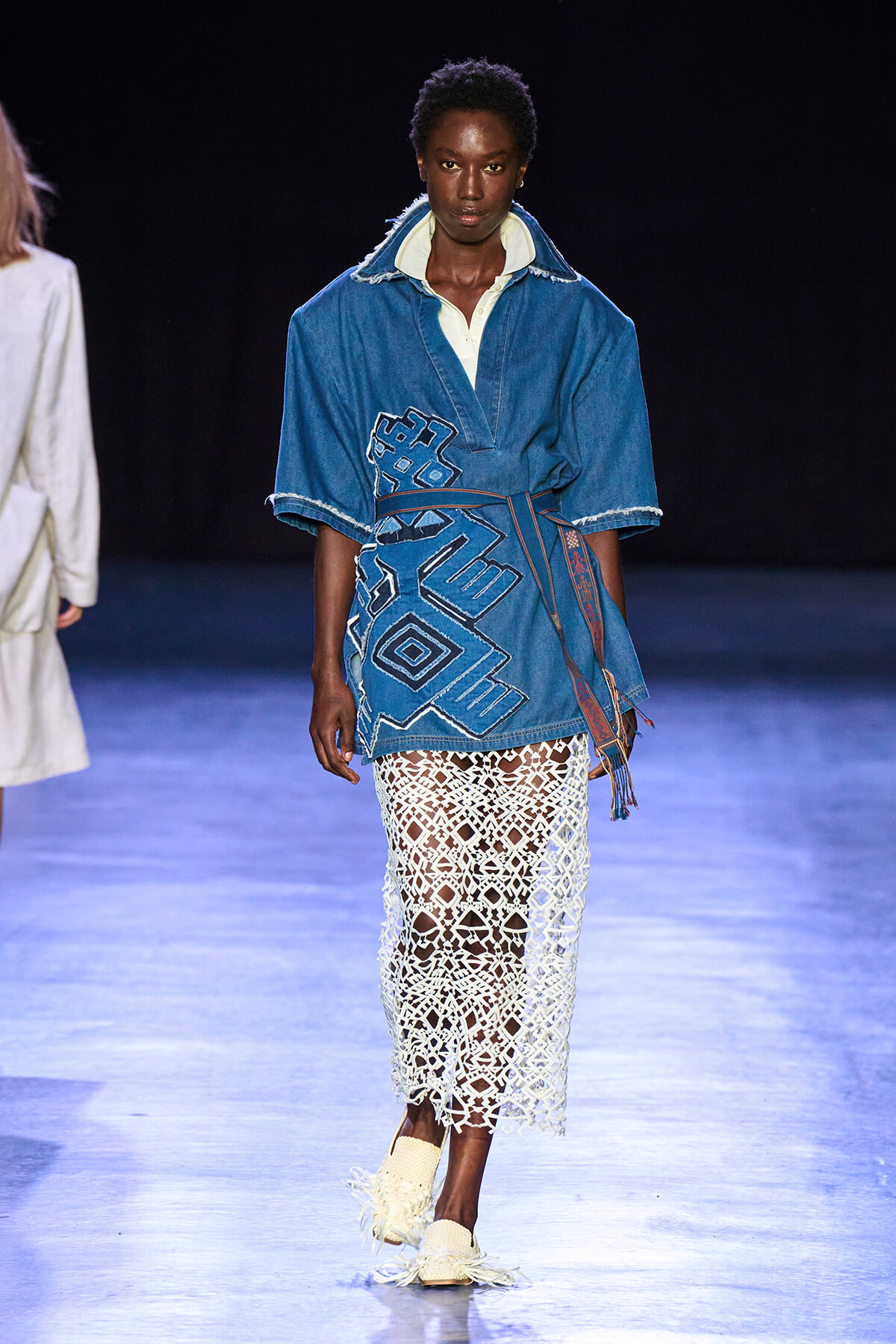 Runway model wears a blue denim tunic with geometric embroidery, paired with a white crochet skirt and light woven shoes.
