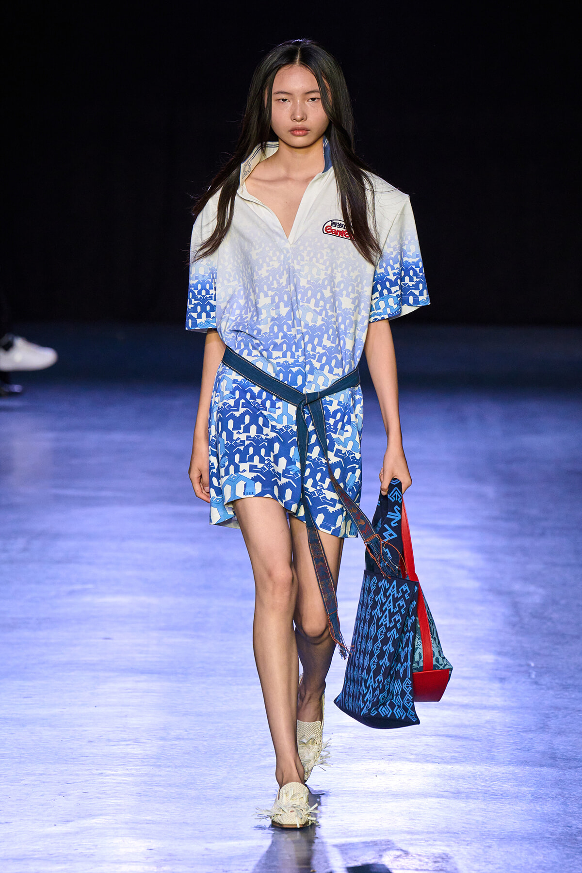 Model walks down a runway in a blue-and-white patterned dress with a dark belt and matching tote bag.