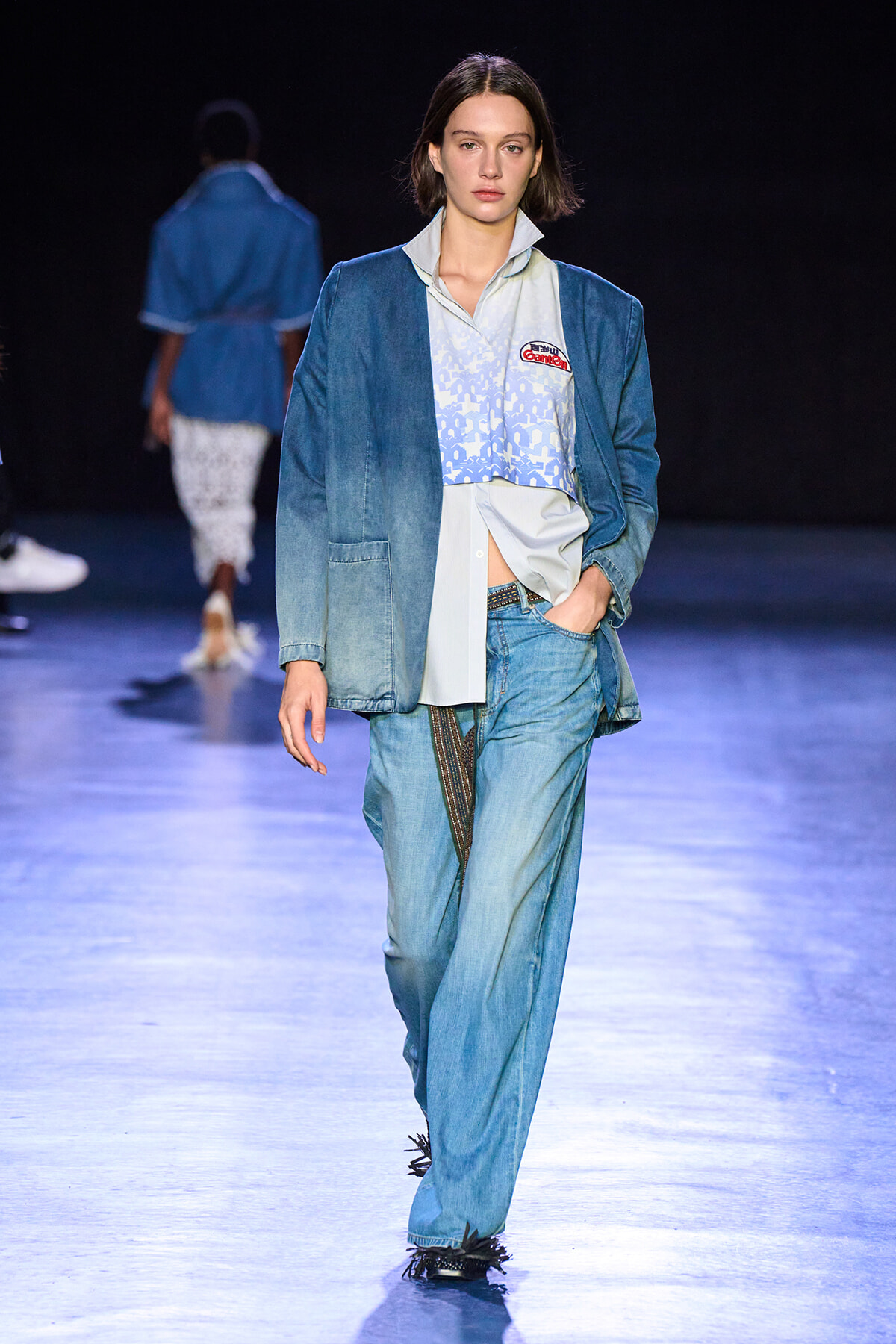 Female model walking a fashion runway in a blue denim blazer and matching light-wash jeans, with a patterned light-blue shirt and patch on chest.