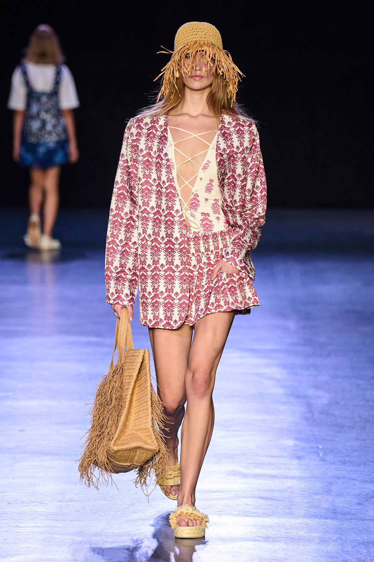 Runway model in a red-printed cream suit with a fringe straw hat, lattice-front top, and a woven straw bag on a dark catwalk.
