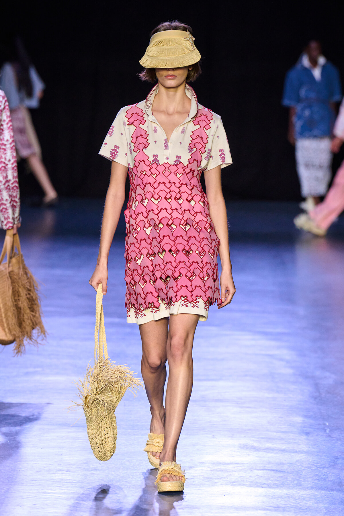 Model walks a runway wearing a cream-to-pink lace-patterned sundress with a layered geometric lattice design, paired with a straw visor-style hat and woven straw bag.