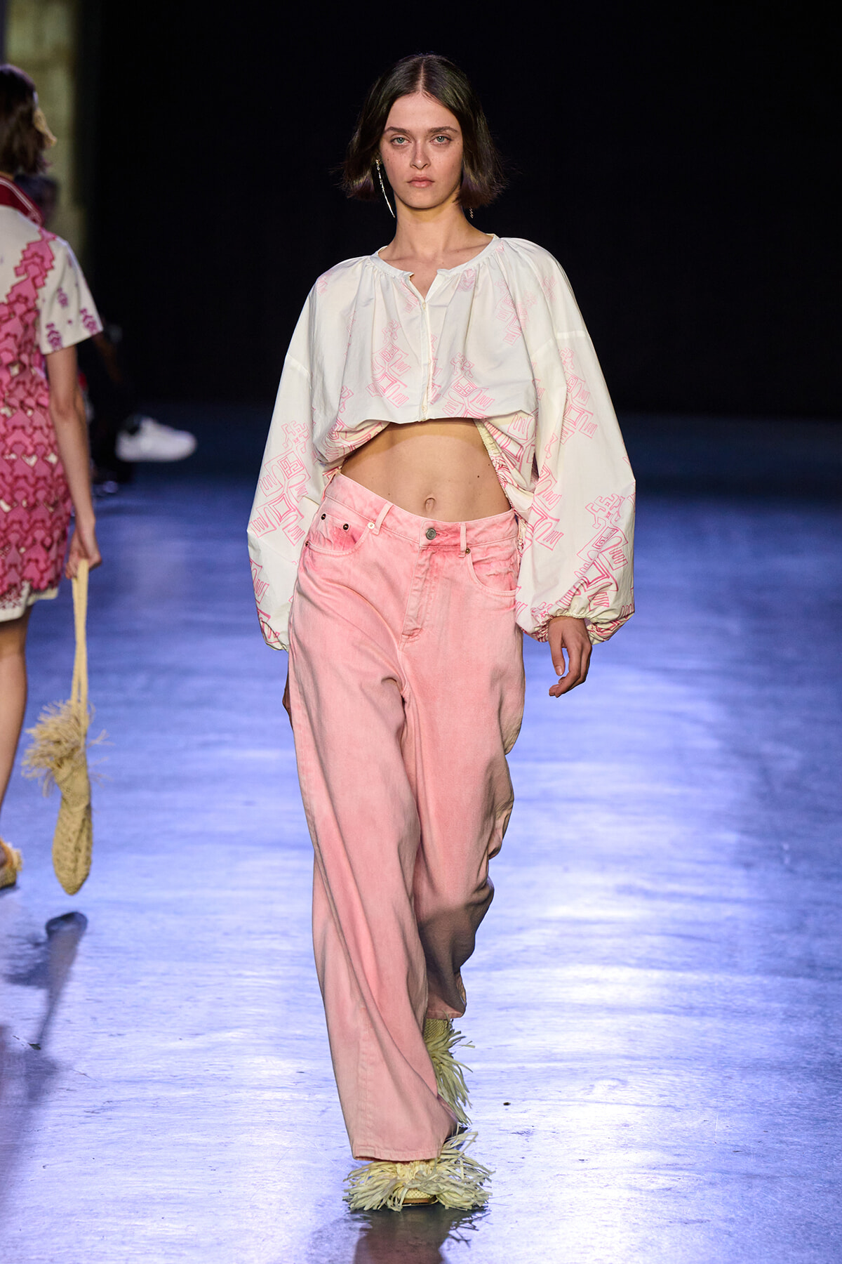 Model walks the runway in a cropped white blouse with pink pattern and high-waisted pink jeans, blue-lit catwalk in background.