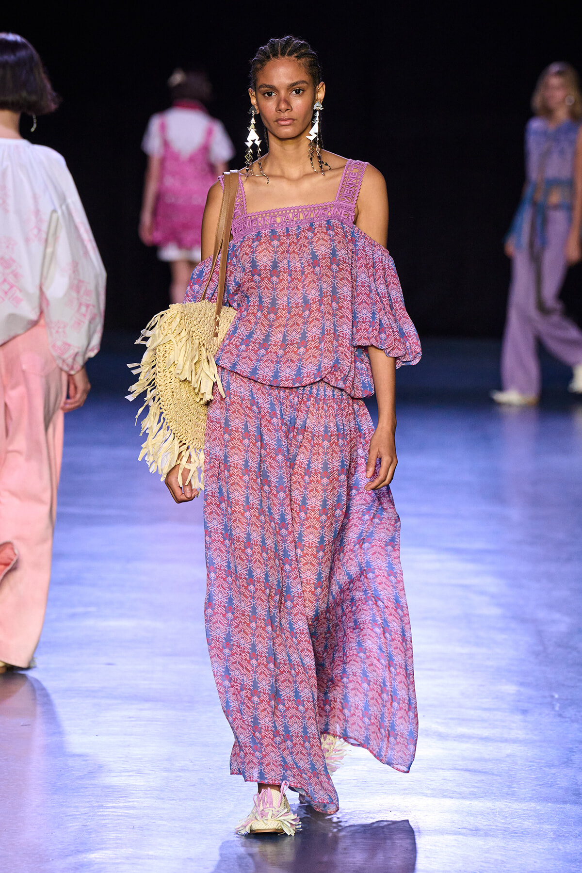 Model walks the runway in a purple-pink geometric print off-shoulder top with matching wide pants, carrying a fringed straw bag and wearing long dangling earrings.