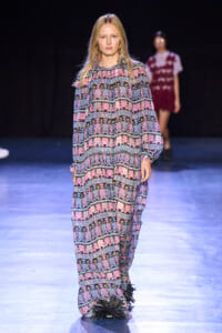 Front-on view of a female model walking a runway in a loose, long-sleeve jumpsuit with pink, purple, blue geometric patterns on a dark background.