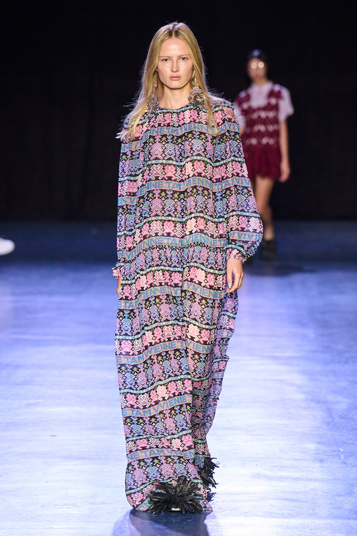 Front-on view of a female model walking a runway in a loose, long-sleeve jumpsuit with pink, purple, blue geometric patterns on a dark background.