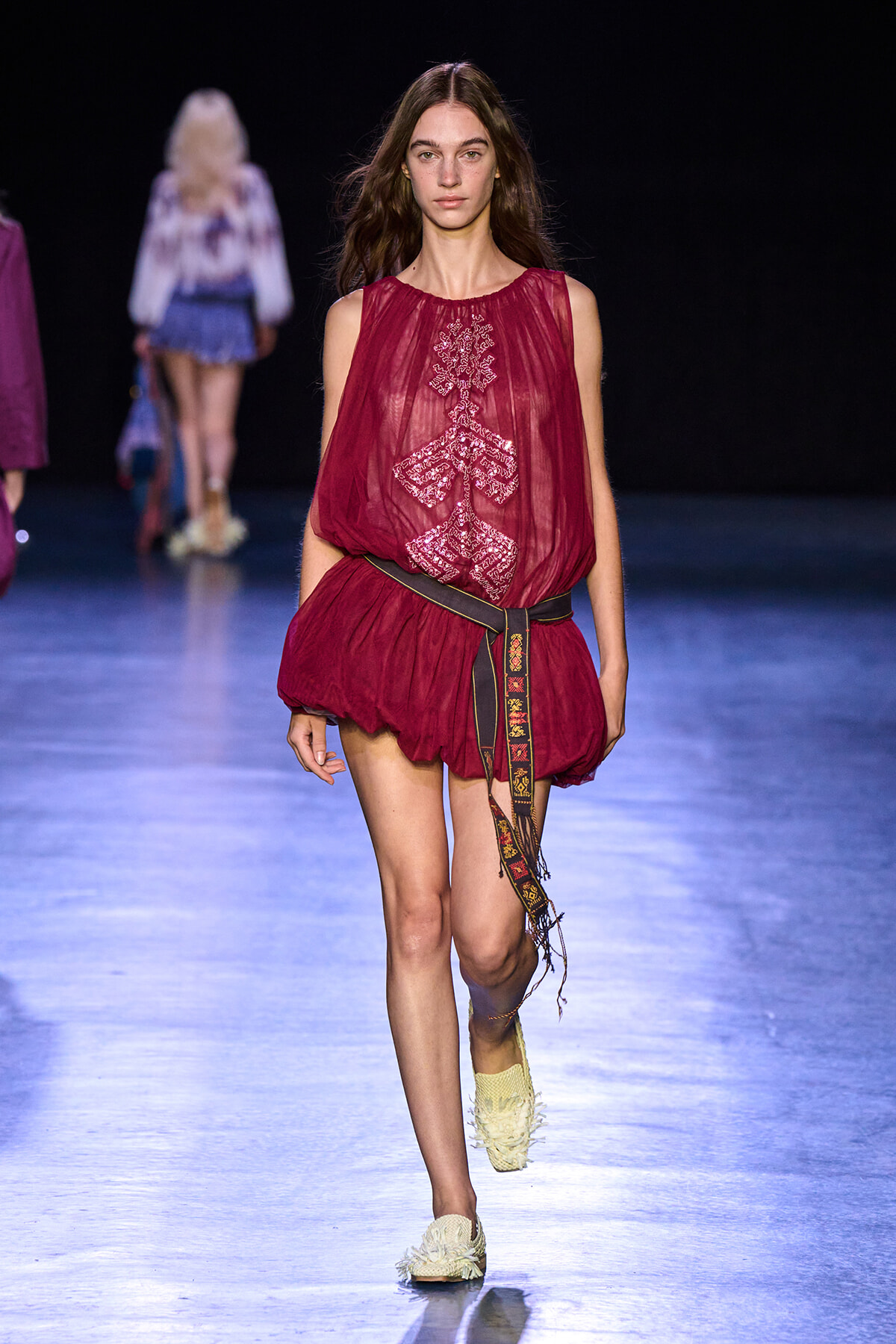 Model walking a runway in a sheer burgundy top with intricate white embroidery and a wide patterned belt.