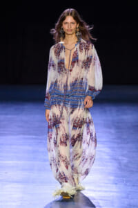 Model walking a runway in a flowing, patterned blue-white-maroon jumpsuit with fringe details and statement earrings on a dark stage.