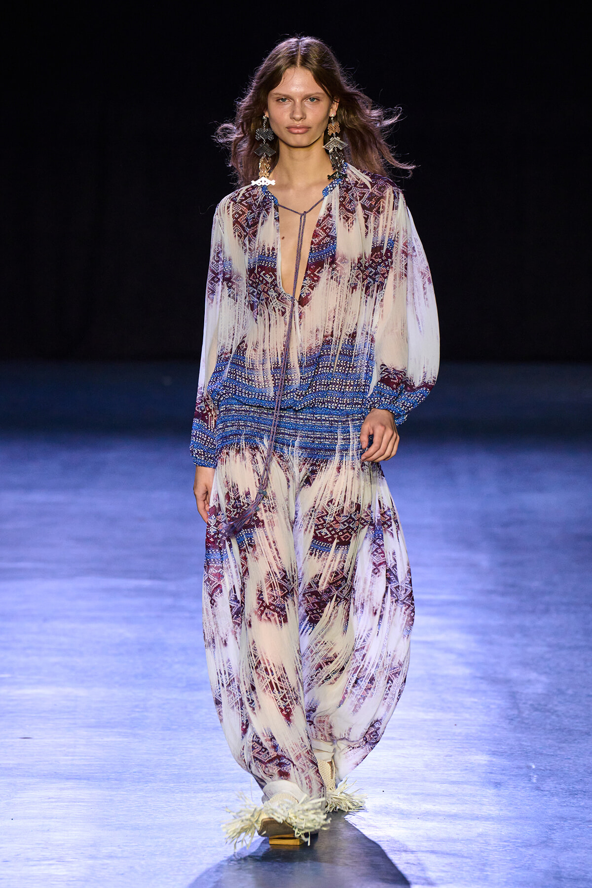 Model walking a runway in a flowing, patterned blue-white-maroon jumpsuit with fringe details and statement earrings on a dark stage.