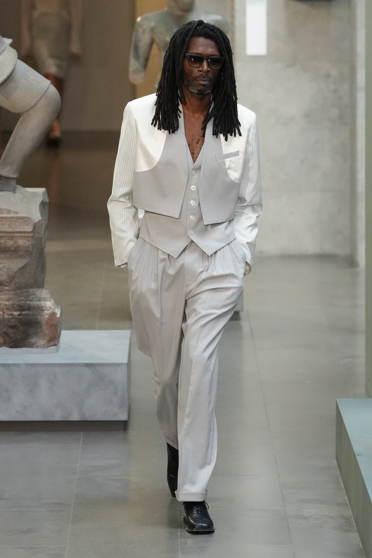 Man with black dreadlocks and sunglasses wearing a light gray three-piece suit walking through a minimalist indoor space.