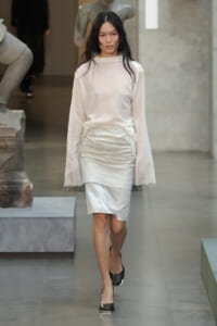 Female model wearing an ivory sheer blouse and crinkled white knee-length skirt, walking down a runway in black heels.