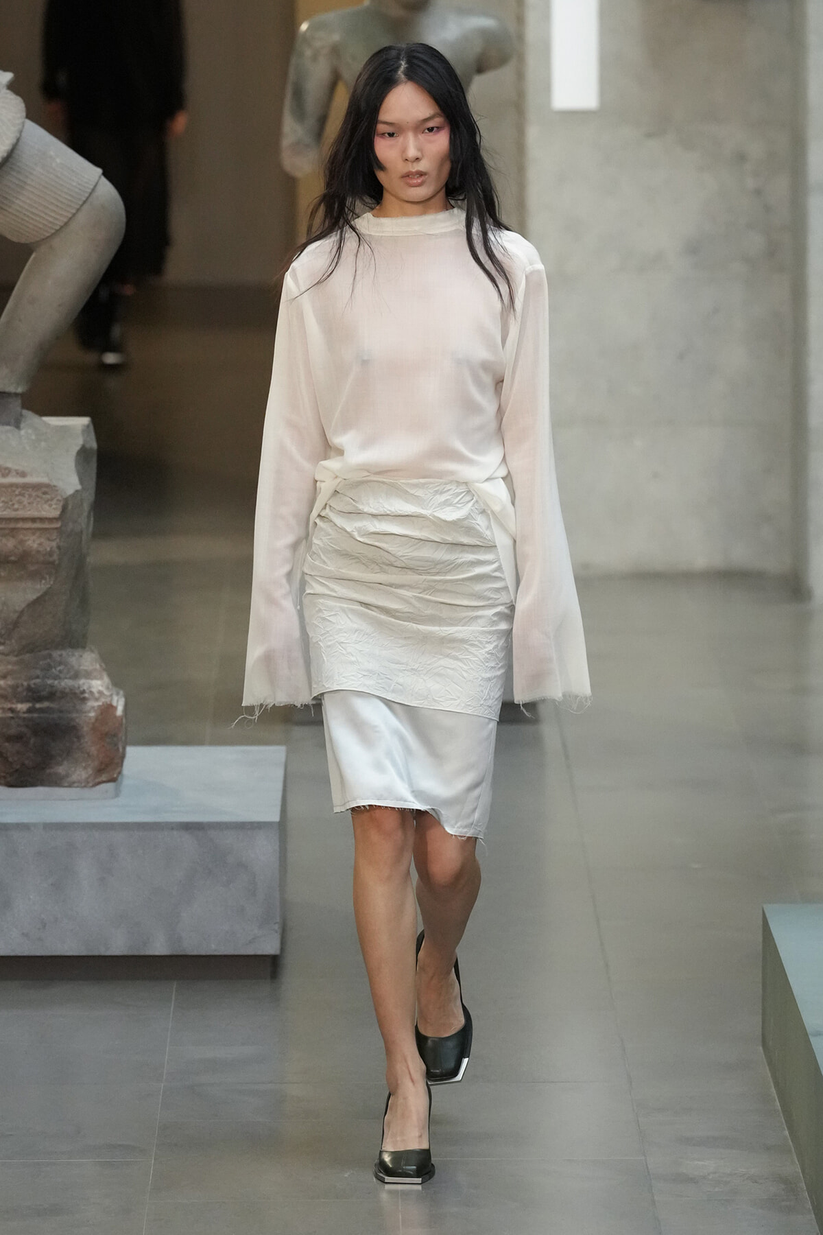 Female model wearing an ivory sheer blouse and crinkled white knee-length skirt, walking down a runway in black heels.