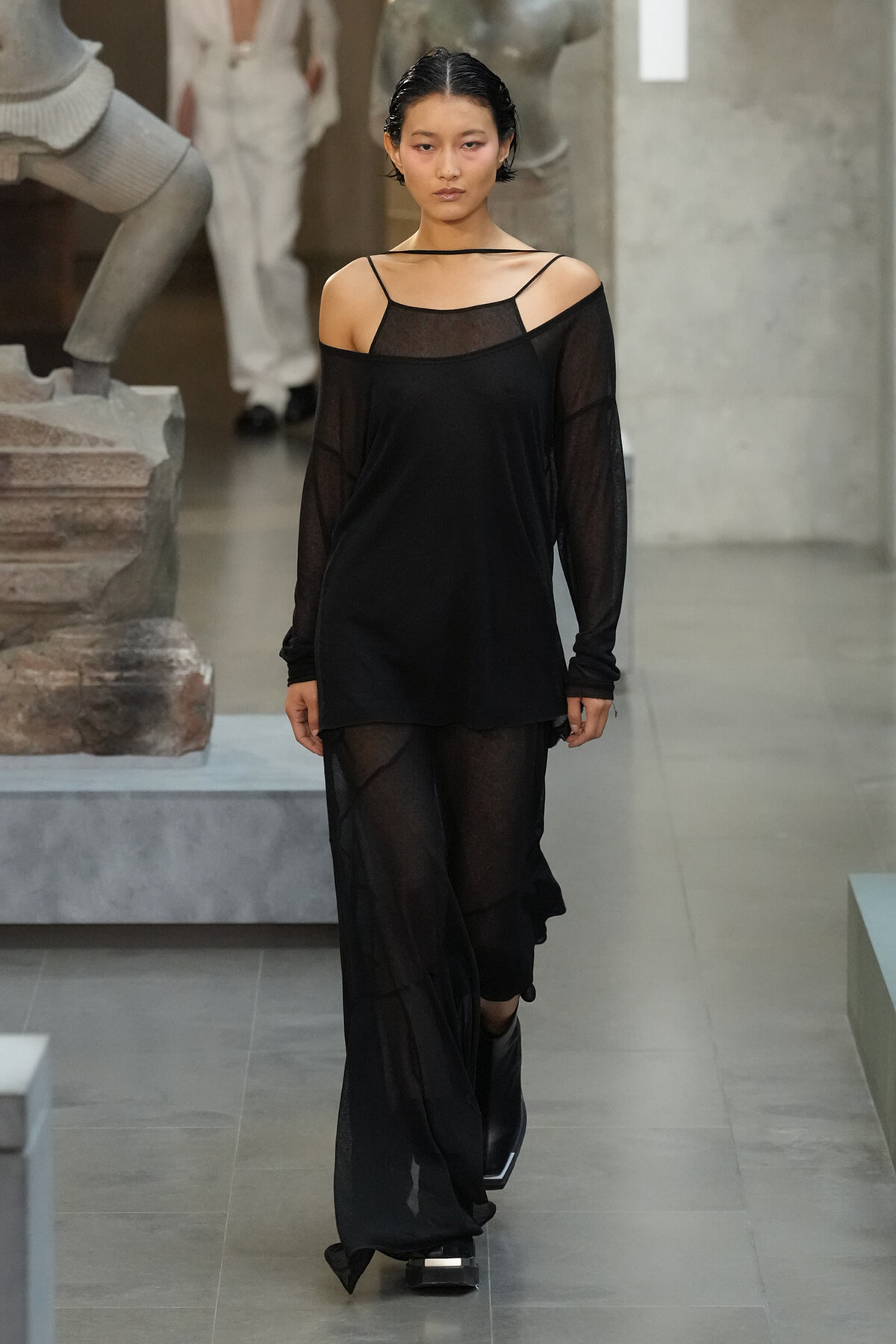 Model walking a runway in an all-black sheer outfit with a loose top and wide-leg pants in an art gallery-like venue.