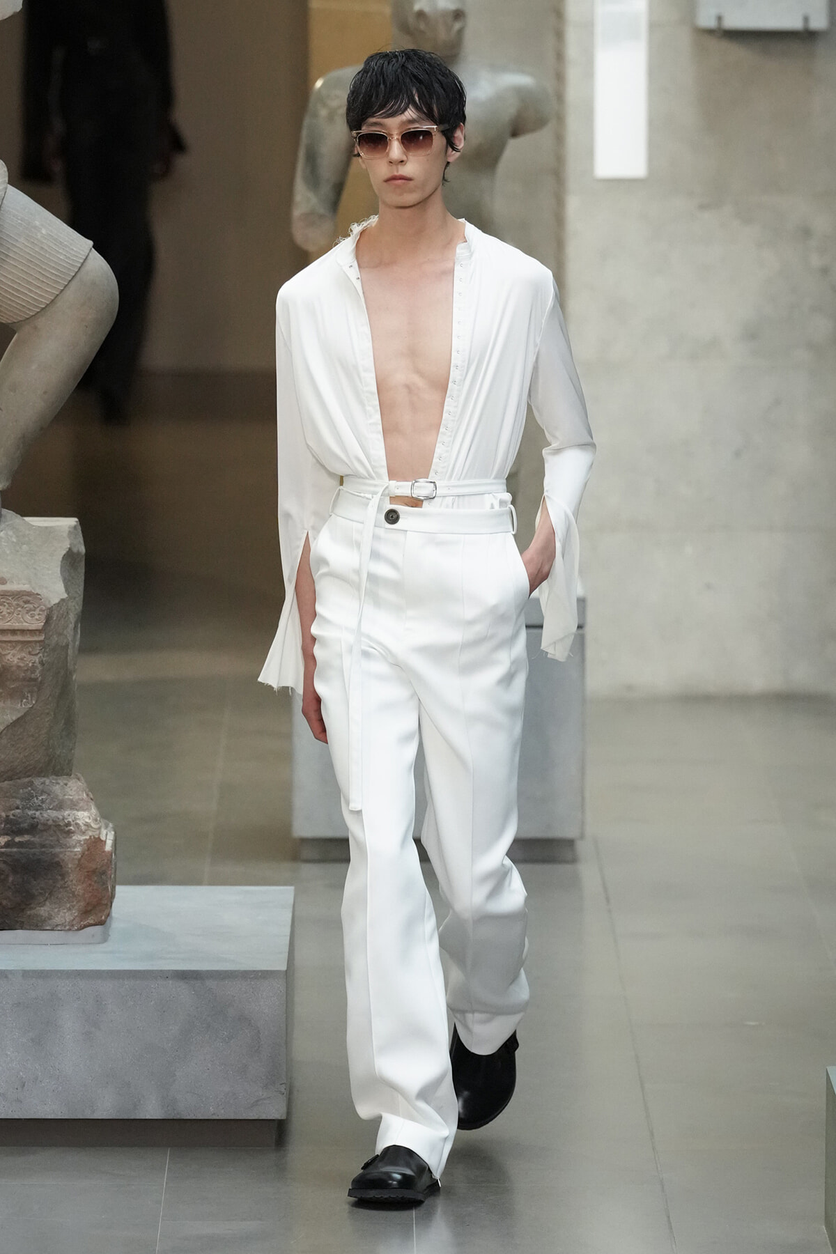 Male model in an all-white outfit with a deep V-neck shirt, belt, and high-waisted pants, wearing sunglasses as he walks.