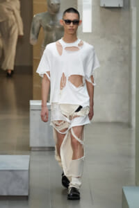 Male model on a runway wearing a white distressed T-shirt with multiple cutouts and ripped beige pants, plus black sunglasses.