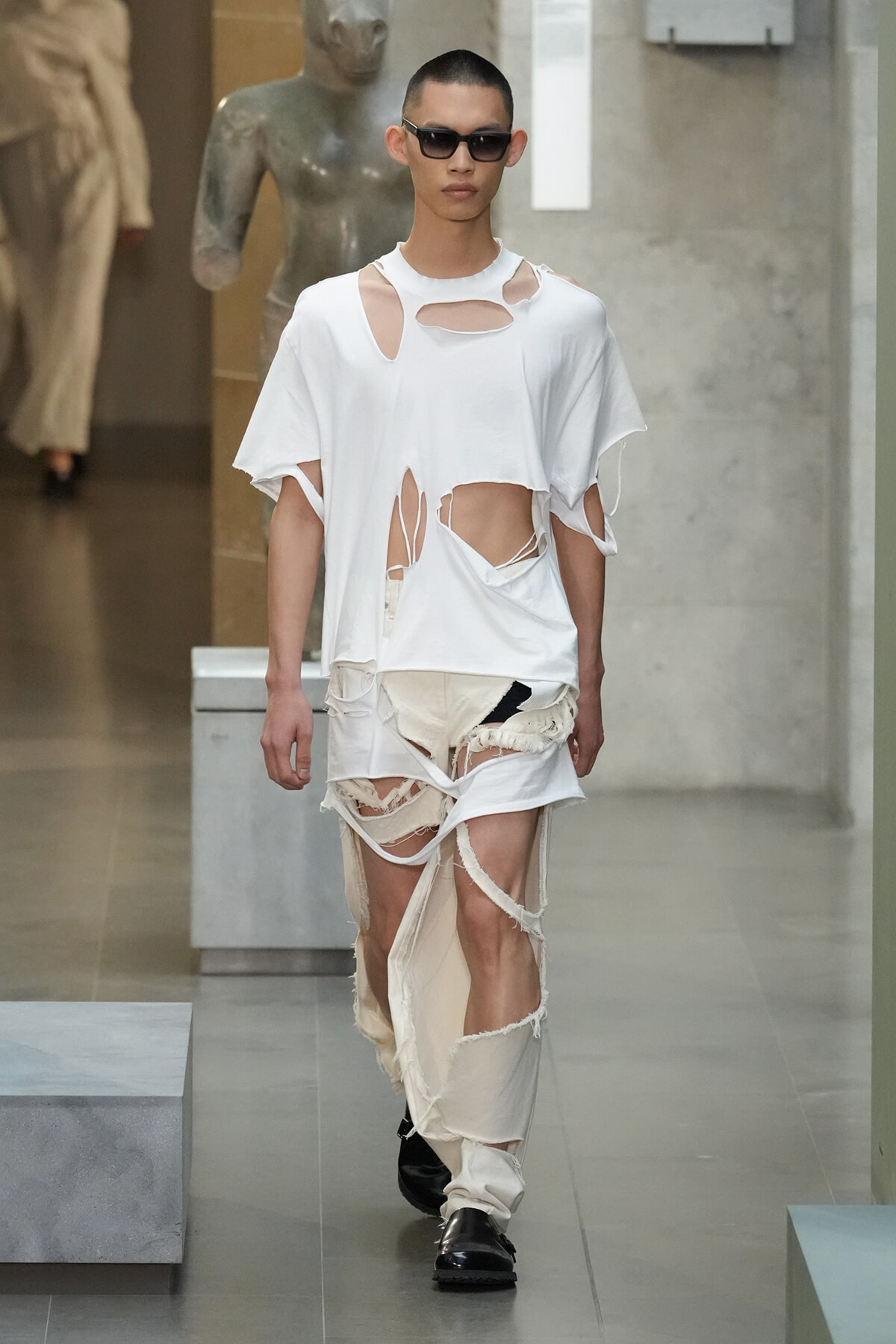 Male model on a runway wearing a white distressed T-shirt with multiple cutouts and ripped beige pants, plus black sunglasses.
