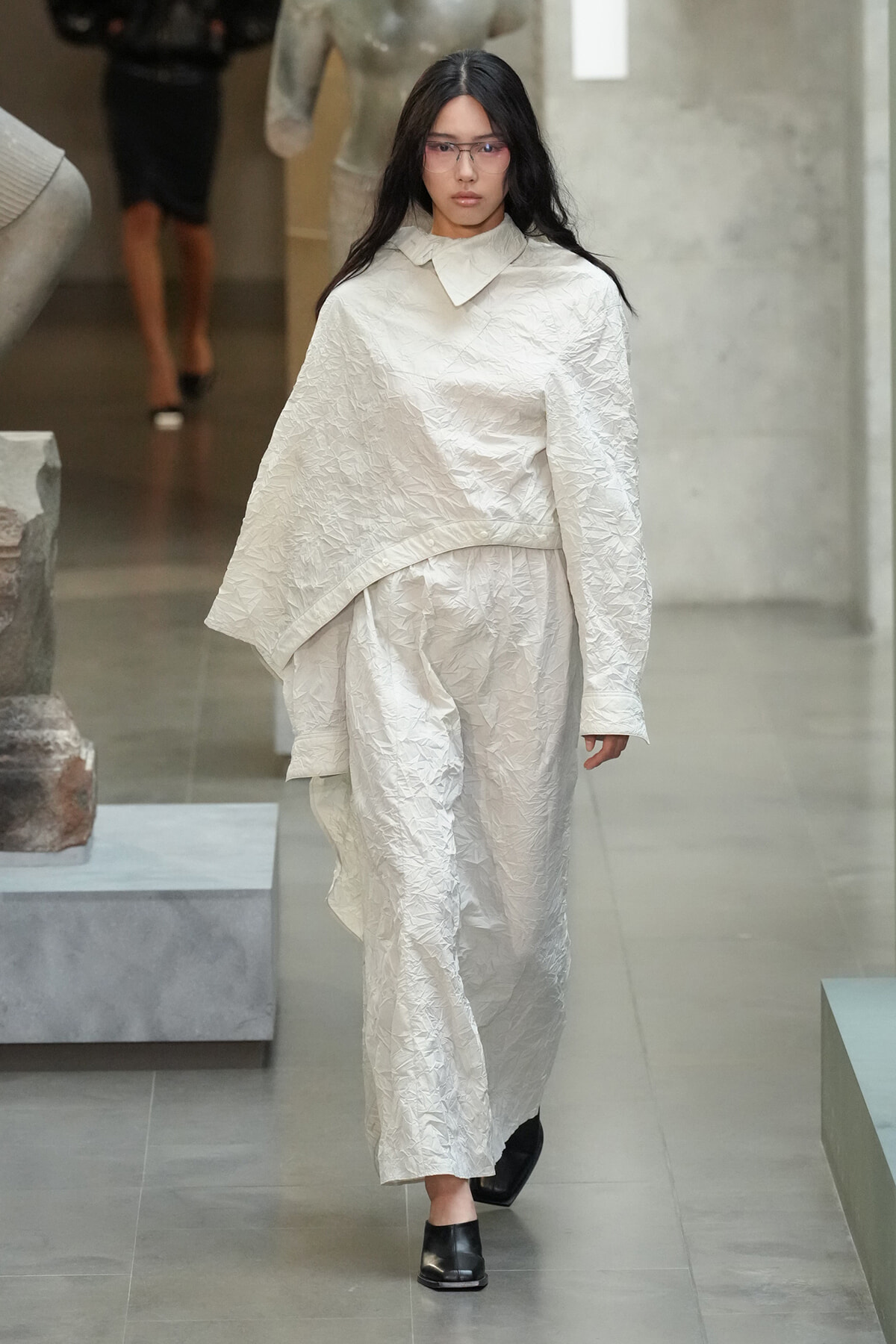 Model walks a runway in an oversized crinkled white outfit with a wide top and matching pants, wearing glasses.