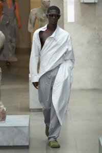 Male model on a runway wearing an oversized white shirt wrapped over light gray pants, sunglasses, and olive shoes.