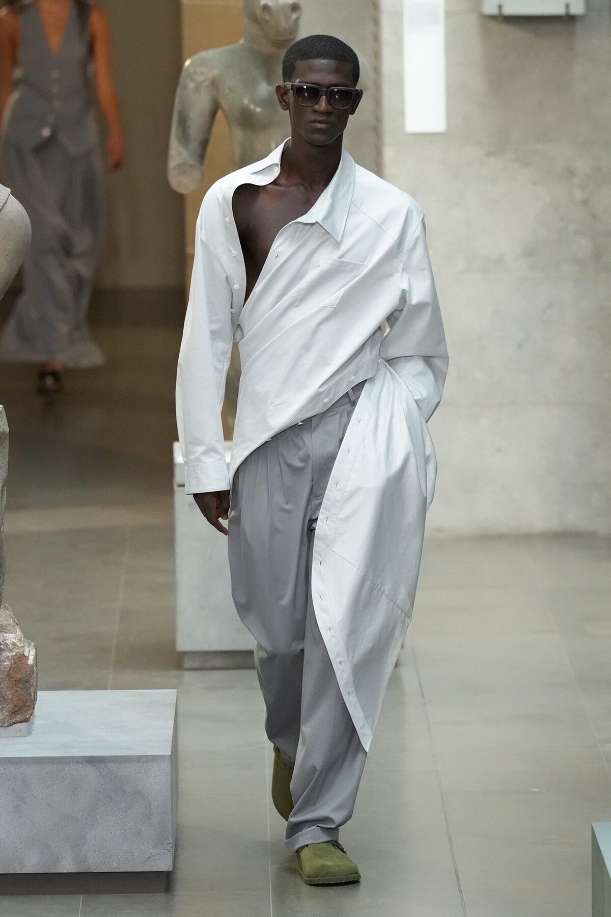 Male model on a runway wearing an oversized white shirt wrapped over light gray pants, sunglasses, and olive shoes.