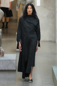 Model wearing a black textured top and pleated wide-leg pants walks through a modern gallery space with sculpture in the background, neutral expression.