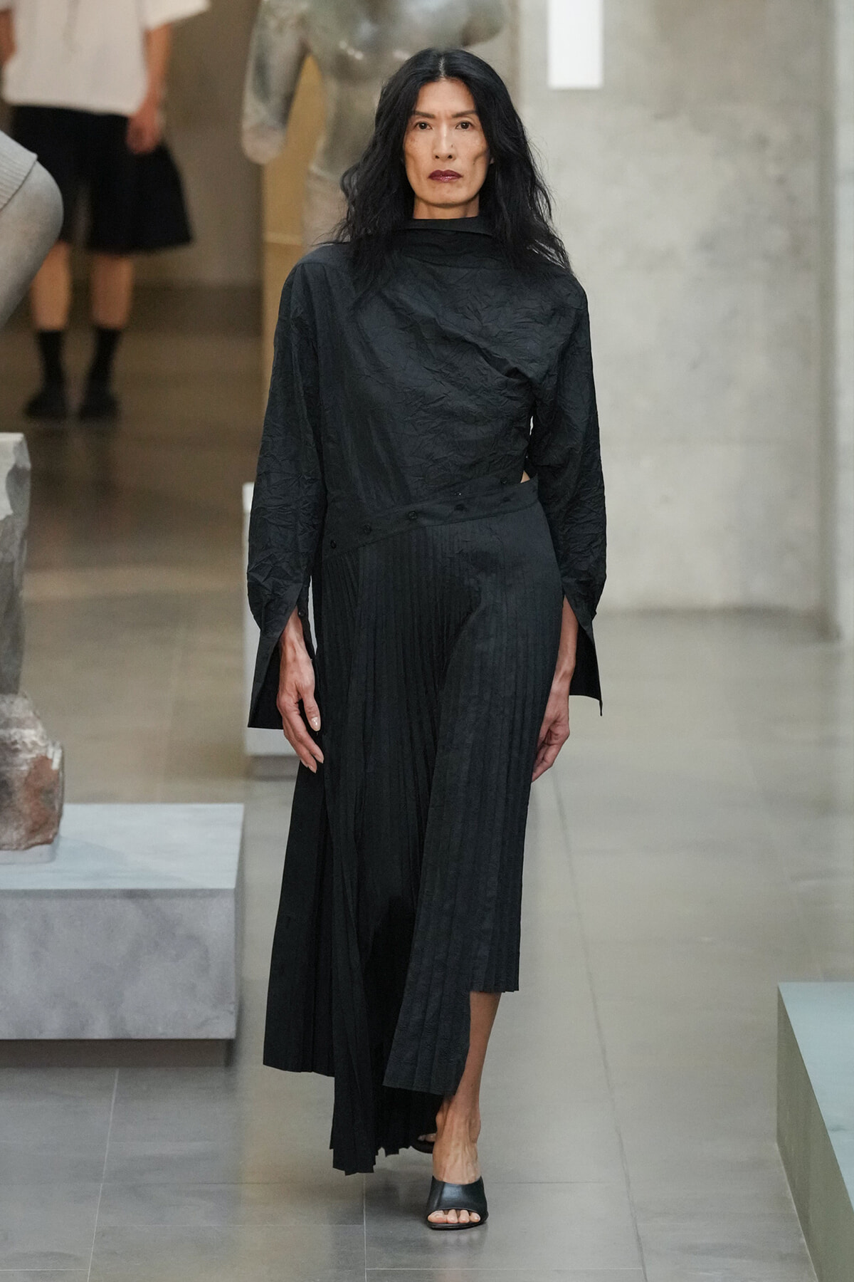 Model wearing a black textured top and pleated wide-leg pants walks through a modern gallery space with sculpture in the background, neutral expression.