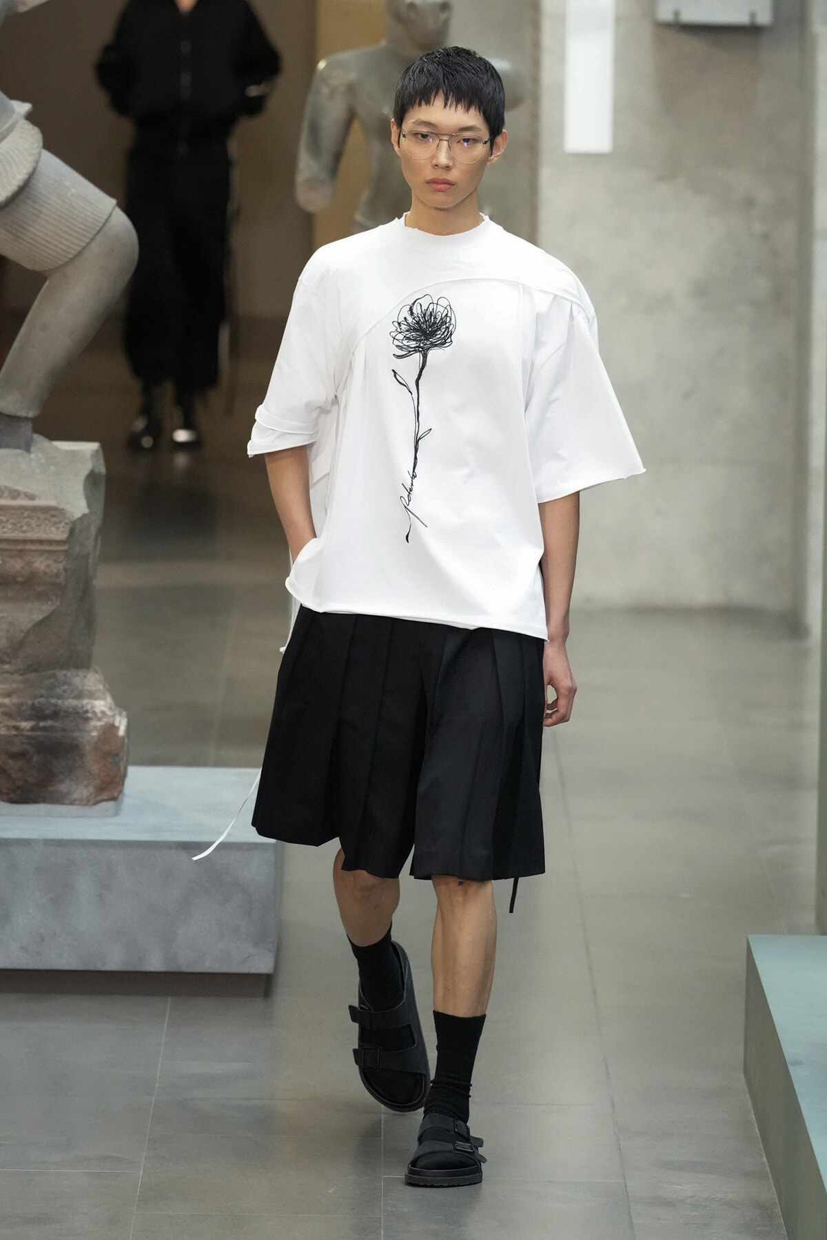 Model walking in a white oversized T‑shirt with a black flower sketch, paired with black pleated shorts and chunky black sandals in a gallery setting.