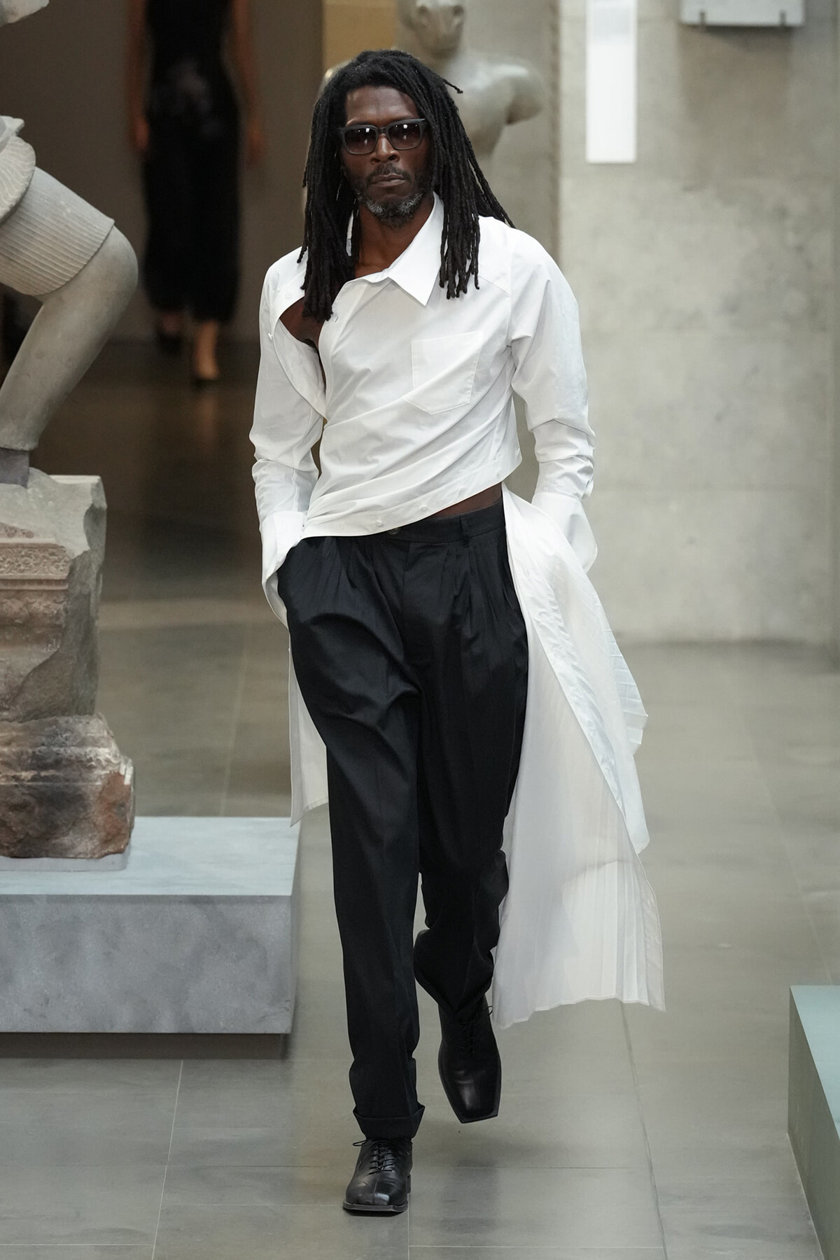 Man with dreadlocks wearing a white shirt and black pants, sunglasses, and a long white coat draped over his shoulders as he walks through a gallery near sculptures,