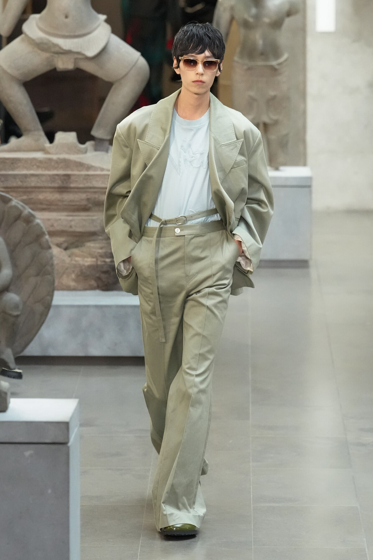 Fashion model in a loose beige suit and sunglasses walking through a museum gallery with stone sculptures in the background.