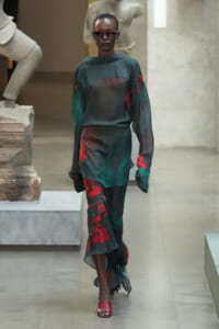 Model walking a runway in a dark green satin blouse with red accents and matching pants, wearing red sunglasses and red sandals.
