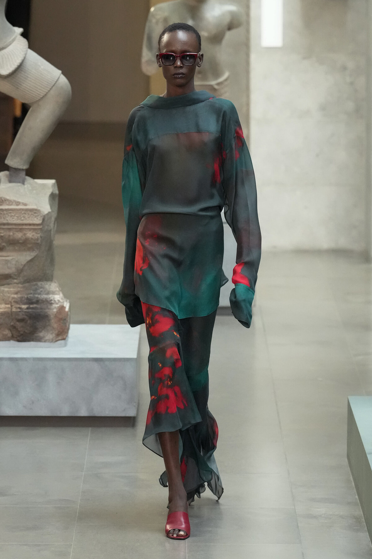 Model walking a runway in a dark green satin blouse with red accents and matching pants, wearing red sunglasses and red sandals.