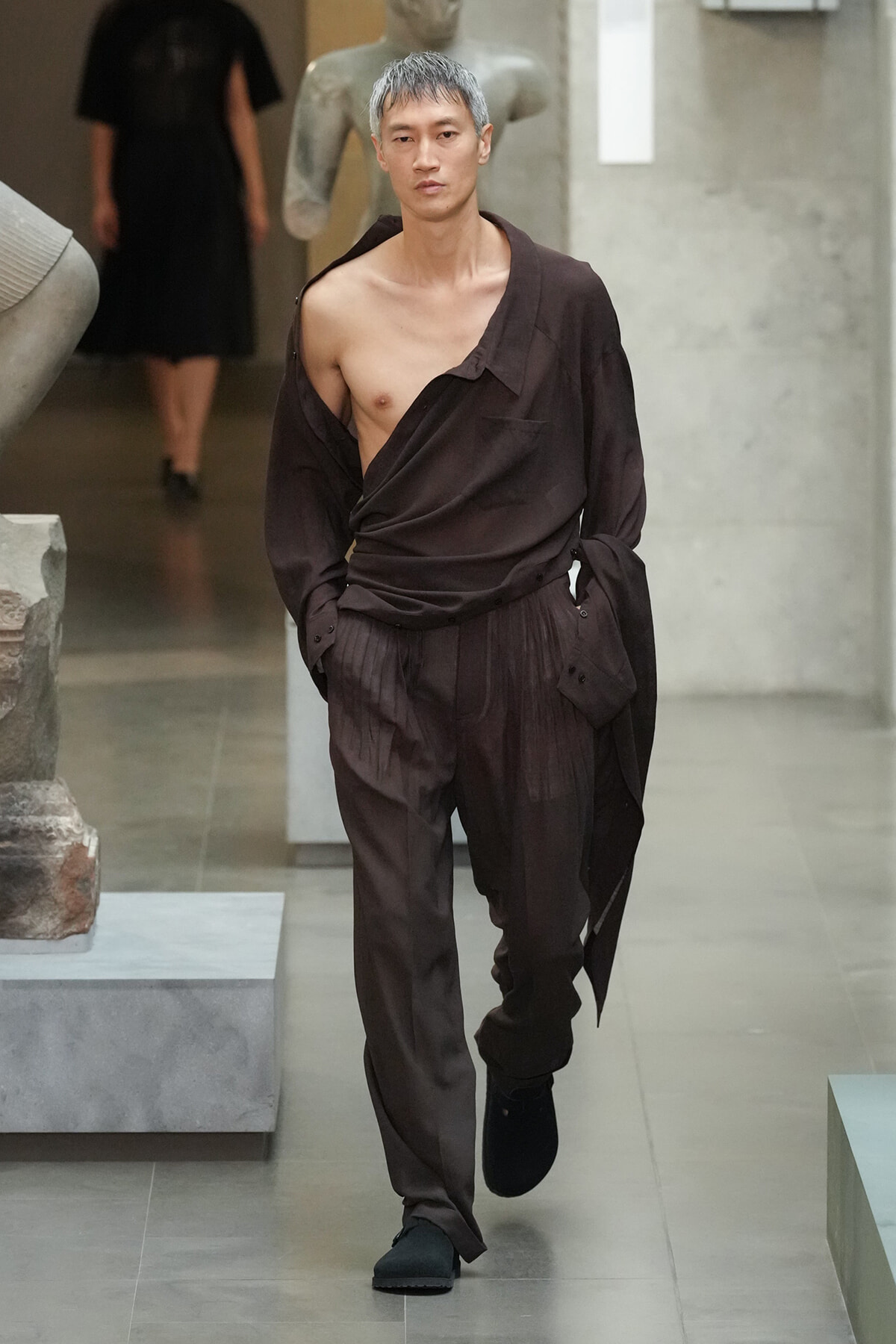 Male fashion model walking a runway in a loose brown outfit with an off-shoulder top exposing one chest, black shoes, in an art-gallery setting.