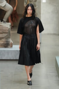 Model walking on a runway in an all-black outfit: sheer black top over a floral design and a pleated midi skirt with black shoes.