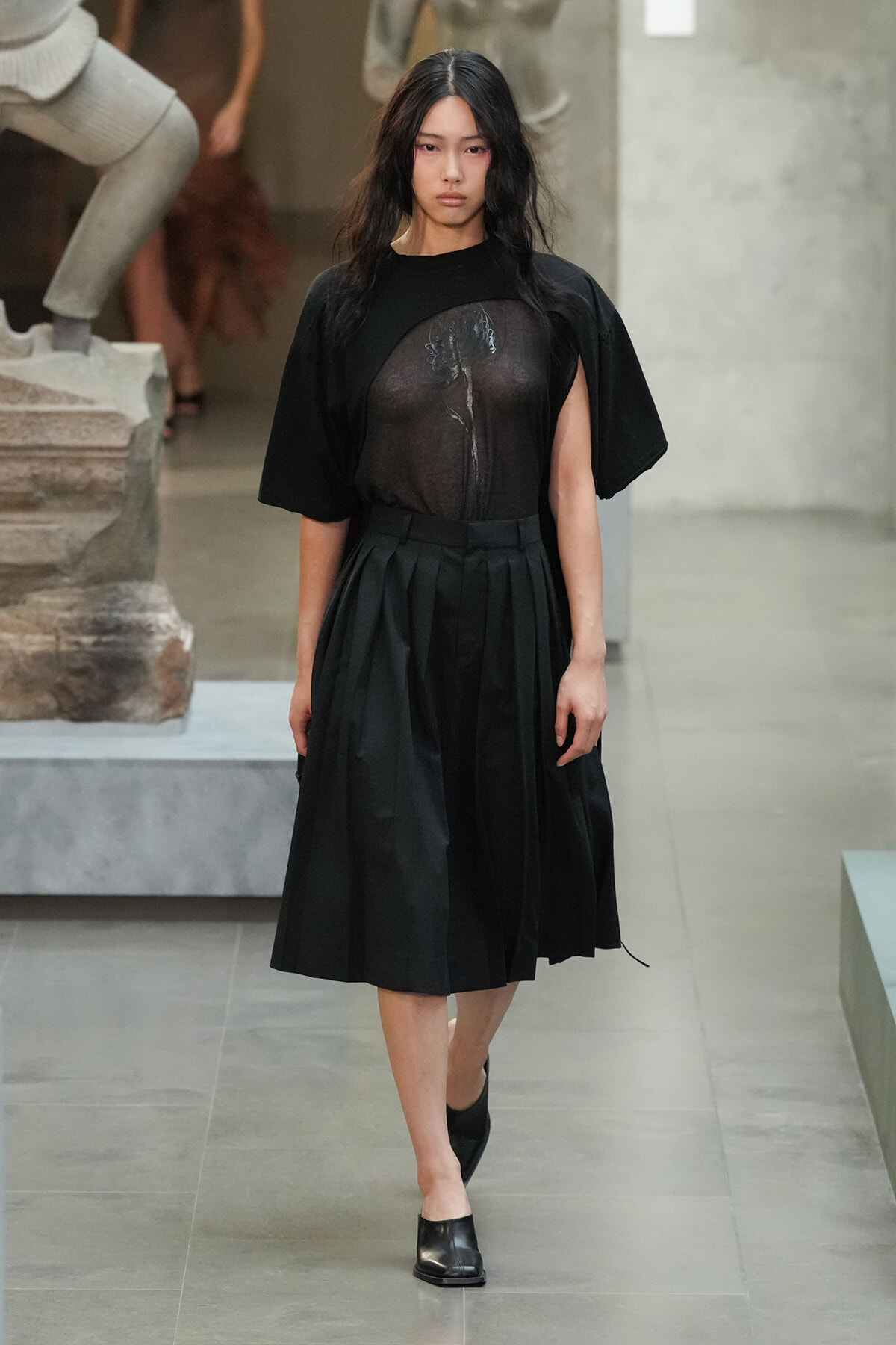 Model walking on a runway in an all-black outfit: sheer black top over a floral design and a pleated midi skirt with black shoes.