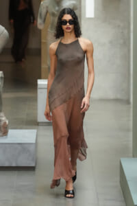 Model walks the runway in a taupe sheer halter dress with an asymmetric hem, paired with black sandals and sunglasses.