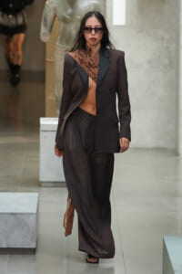 Female model walks a fashion runway wearing an oversized black blazer over a brown draped blouse and wide-leg pleated black pants, with dark sunglasses and open-toe sandals in a minimalist setting.