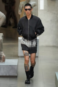 Model walking a runway in a dark bomber jacket, tie-dye shorts, black socks and boots, wearing sunglasses in a gallery setting with sculptures in the background, tattoos visible on leg.